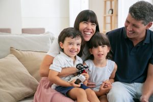 Cheerful funny parents and two kids watching funny movie Cheerful funny parents and two kids watching funny movie at home, sitting on couch in living room and looking away and laughing. Medium shot. Leisure time with family or television concept